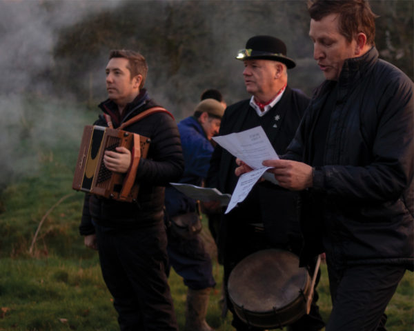 Colour photograph of three white men at a wassail. One holds an accordion, one has a drum around his waist, and one holds some sheets of paper