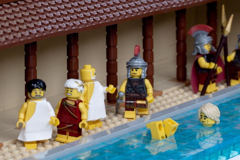 Lego Roman figures at a bath house. There are two figures in togas and a centurion. A pair of feet are sticking up out of the water