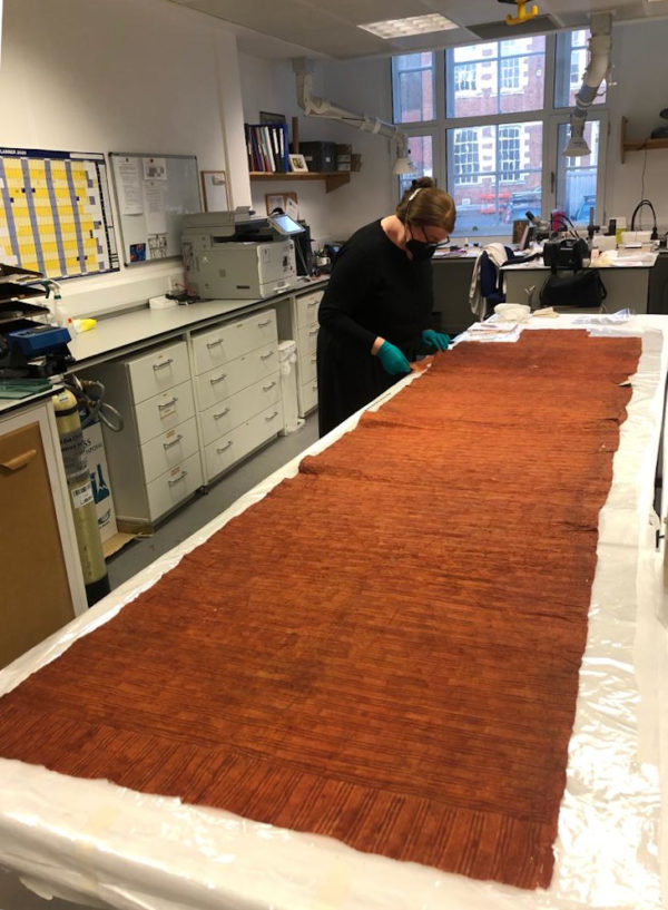 The unrolled barkcloth is on a table in RAMM's conservation lab. A conservator, wearing green gloves, is studying it