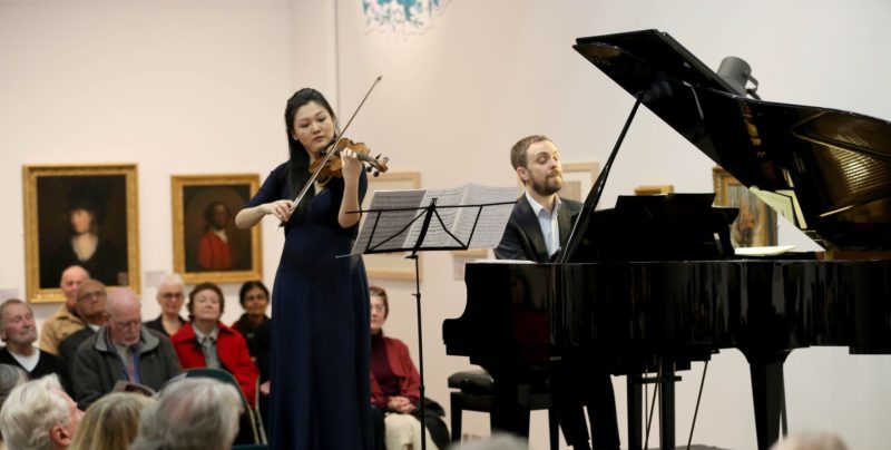 A group of people listen to a woman playing the violin and a man on the piano