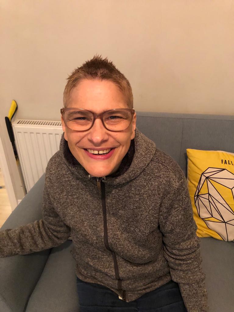 A smiling woman with short fair hair, seated on a grey sofa, wearing glasses and a grey hoodie
