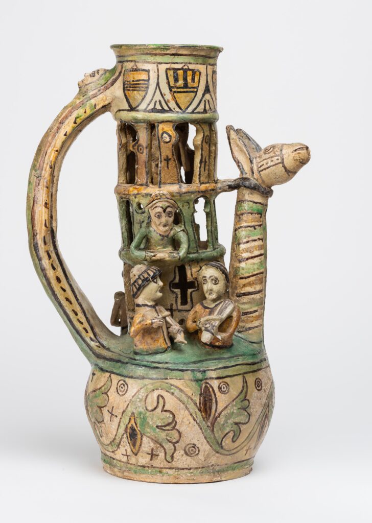 An intricate medieval puzzle jug. The colour is mostly cream or beige, with green and yellow accents. Small figurines are set into the neck of the jug, including a woman and man. The spout takes the form of a giraffe or other long necked animal.