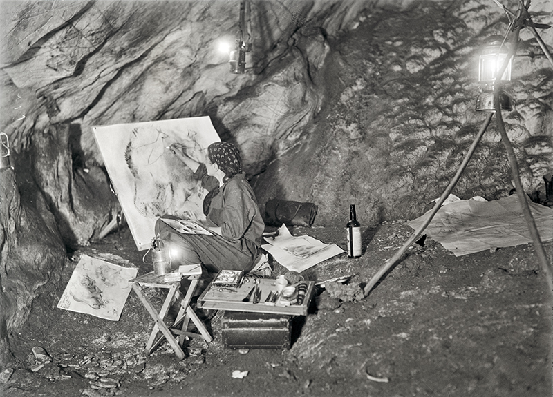 A black and white image of a woman, Elizabeth Pauli, painting in a cave. She wears a head scarf and sits on a wooden stool