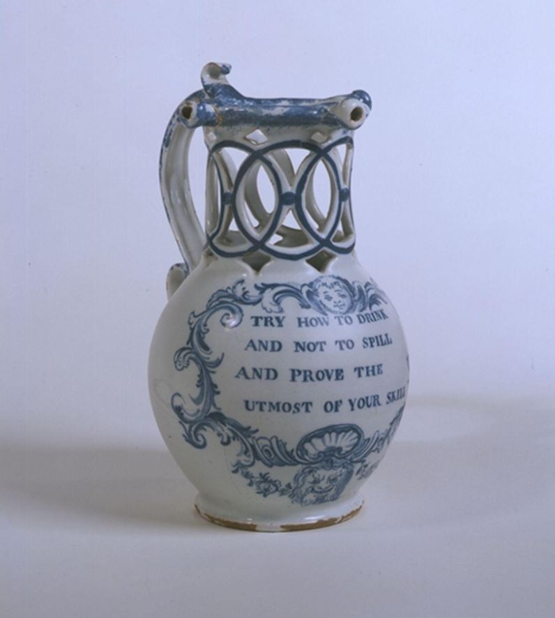 A white puzzle jug with blue decoration and a hollow neck, constructed of interlocking circular patterns. The words 'try how to drink and not to spill to prove the utmost of your skill' are stamped on it