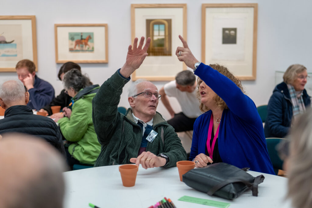 an older gentleman and a woman in a pink lanyard seated at a table in a gallery at RAMM, both pointing at something above them