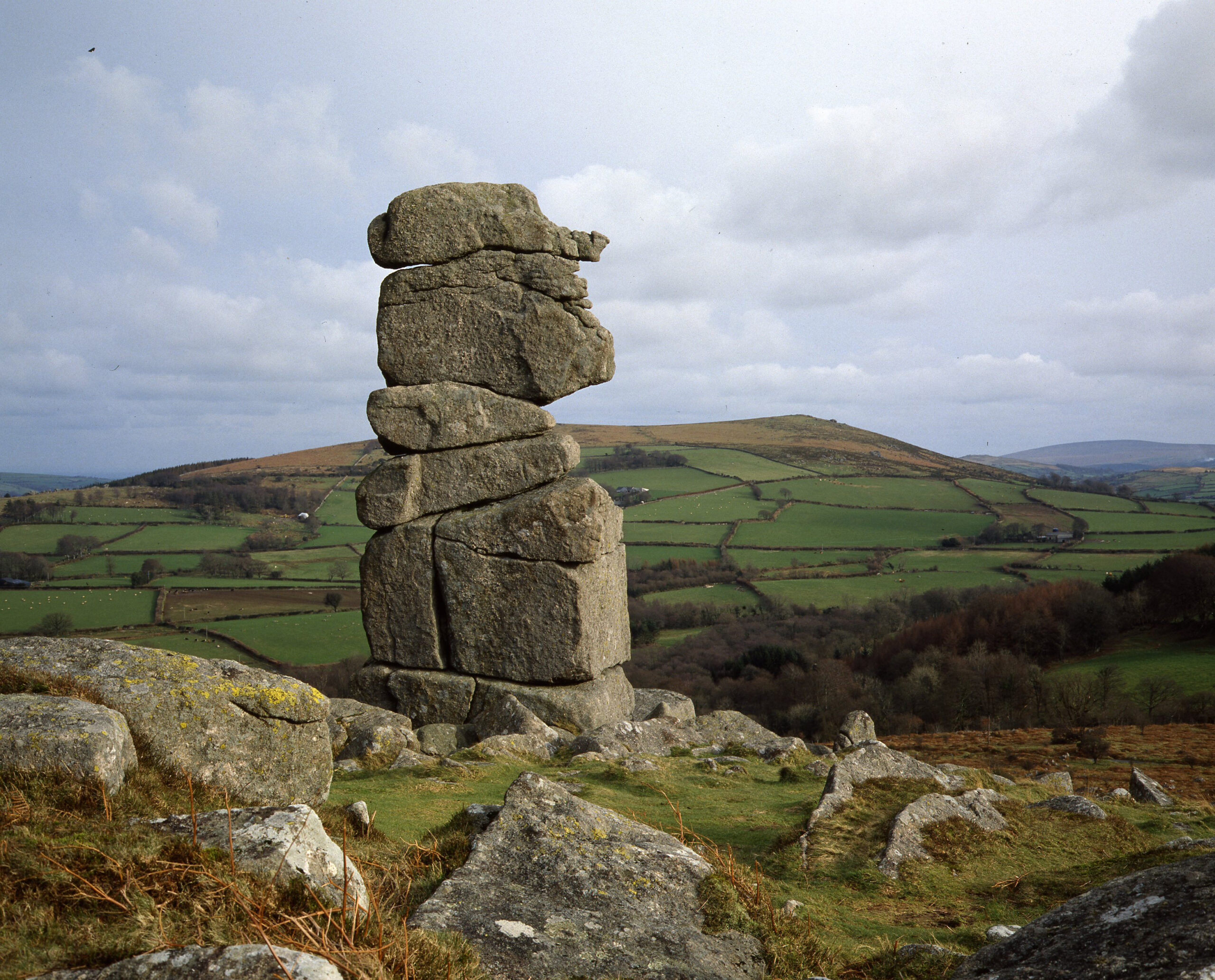 An ancient rocky formation that looks like a body and a head, known as Bowerman's Nose stands proud just off centre left of a photo of the Manaton landscape. Fields can be seen in the distance and the sky is cloudy.