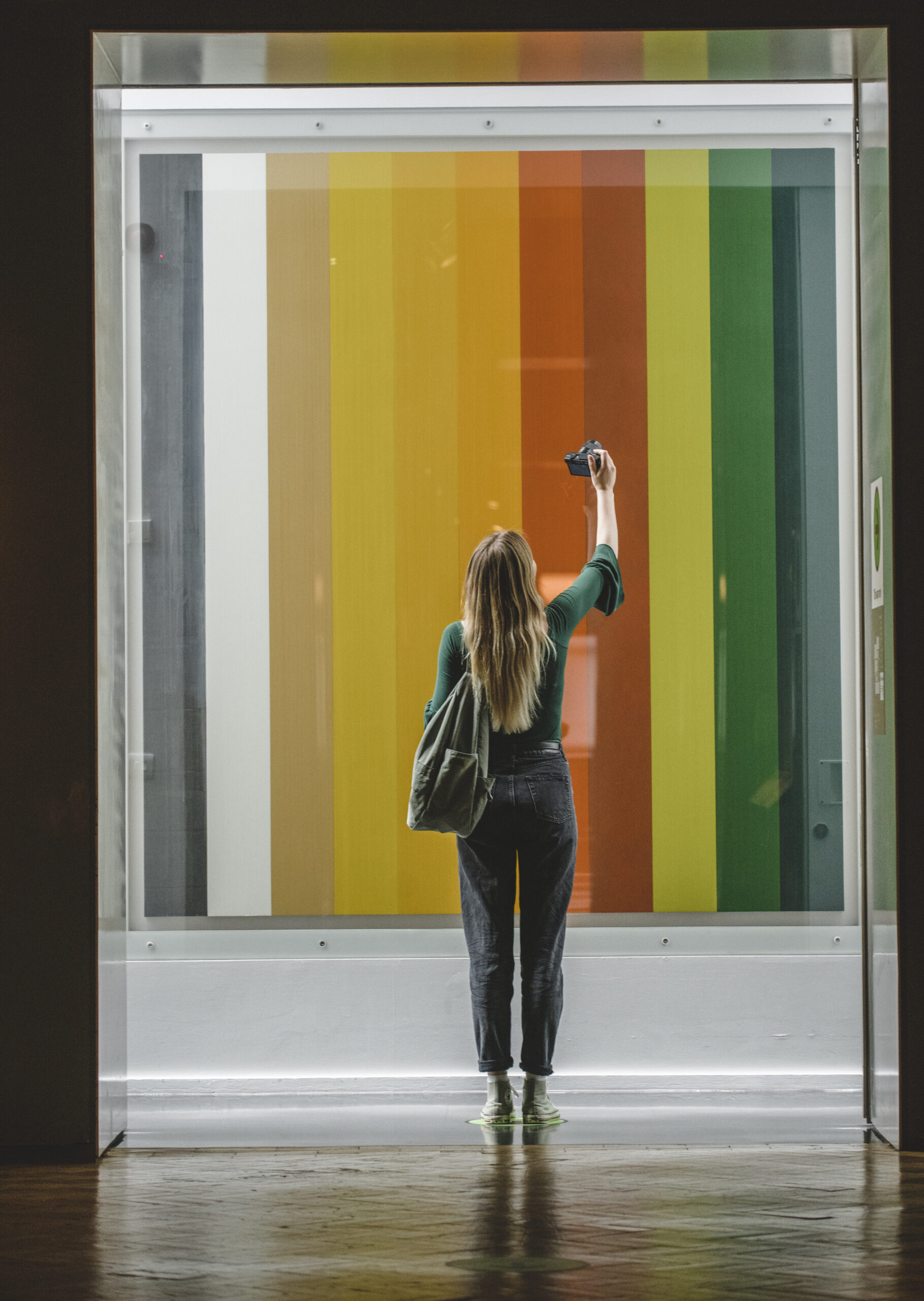 A young woman stands in one of the museum doorways with her back to the camera. She is taking a selfie of herself. In front of her is a contemporary artwork made up of stripes of coloured paint.