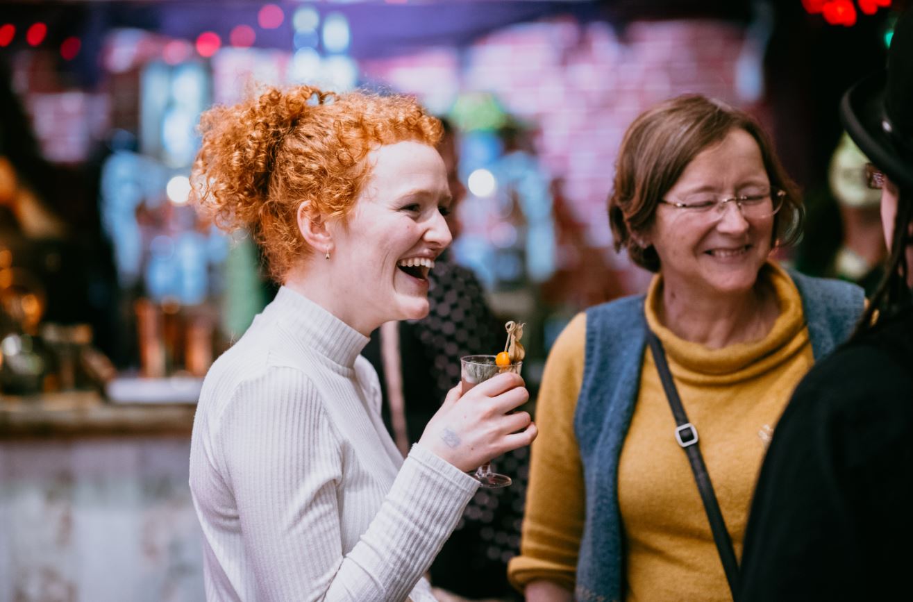 a woman with red hair holding a drink in the museum, with another woman in a yellow top standing next to her. they are engaged in conversation with an unseen third person, and smiling