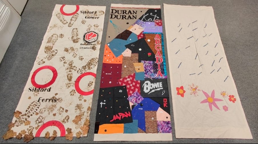 3 vertical fabric banners laid on the floor. One is covered in footprints and mud. ANother has colourful fabric and the words 'duran duran' 'kate bush' 'bowie' and 'Japan'. Another is pale and covered in line marks
