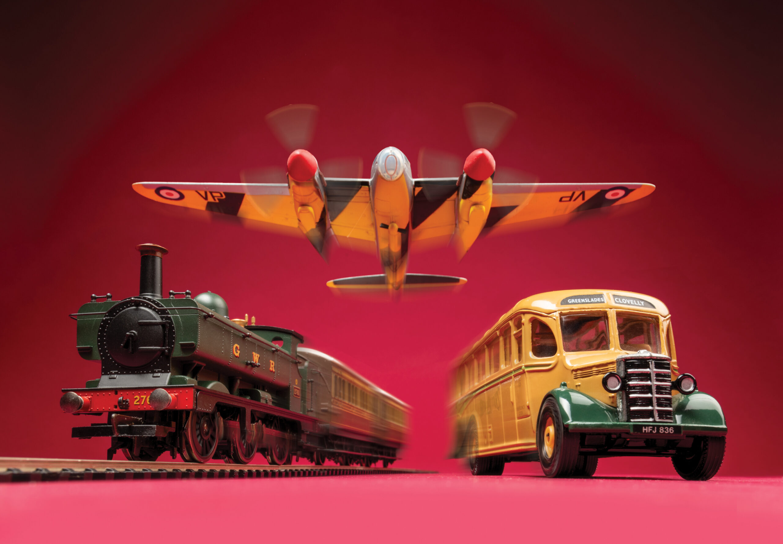 a toy train and bus on a red surface, with a toy plane hanging above them. the background is also red