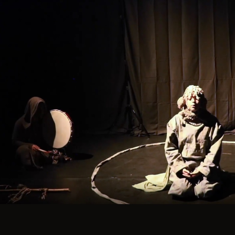 Ashanti Hare, River that never rests 2024, still from a video: a person kneeling in a circle drawn on the floor, against a black backdrop. to the left of the photo a hooded figure holds a large tambourine