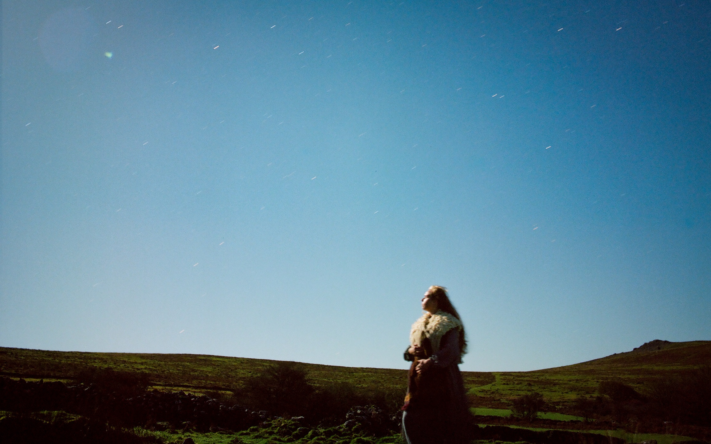 a long exposure photograph of a woman standing on a moor, under a sky full of stars
