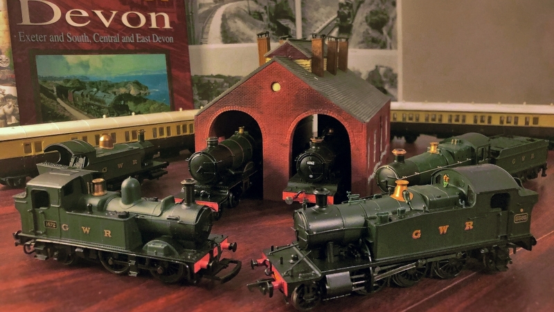 4 model steam trains on a table, two of which are peeking out from inside a model train depot