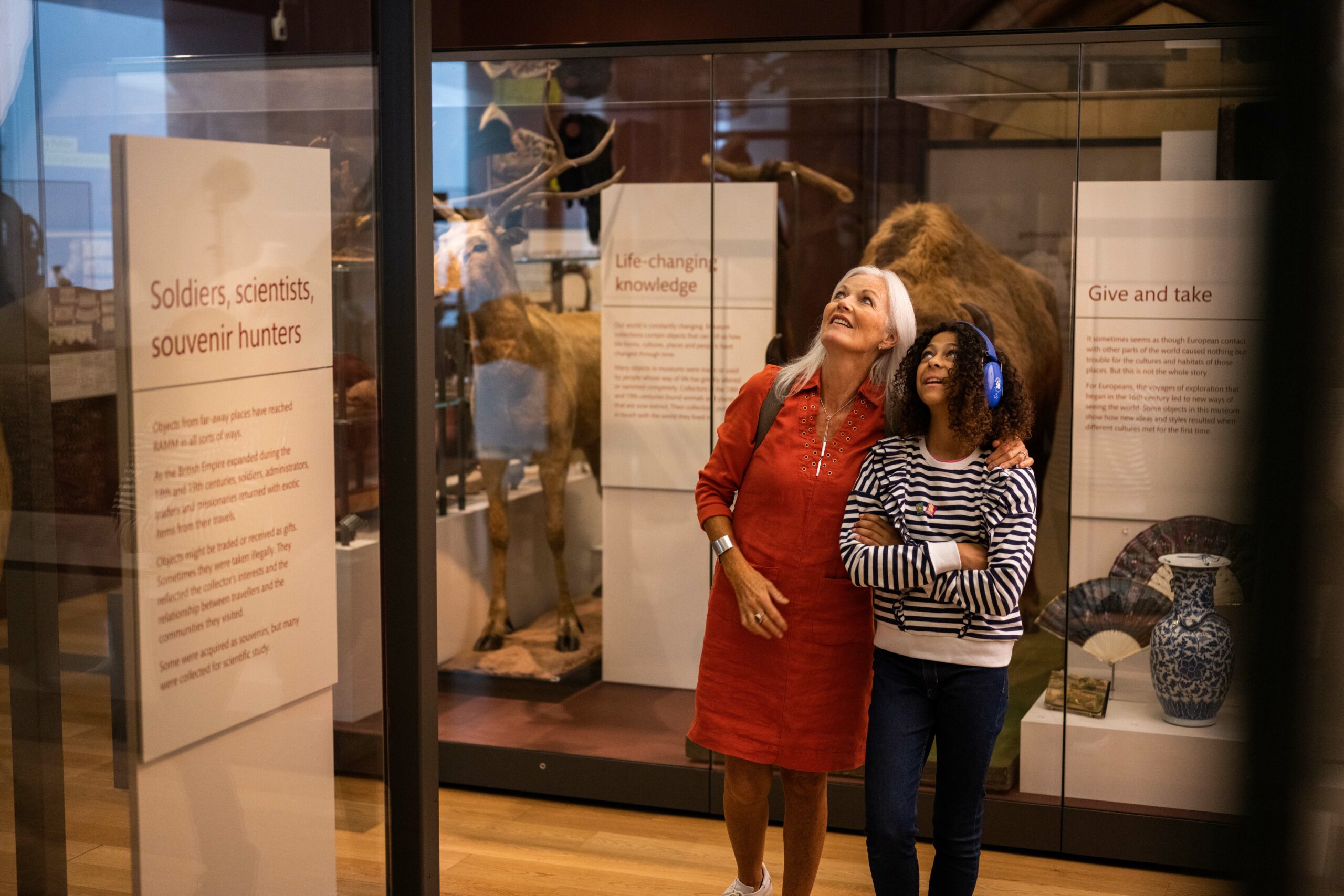 Photo of a lady and a young girl looking at museum cases. The young girl is wearing ear defenders.