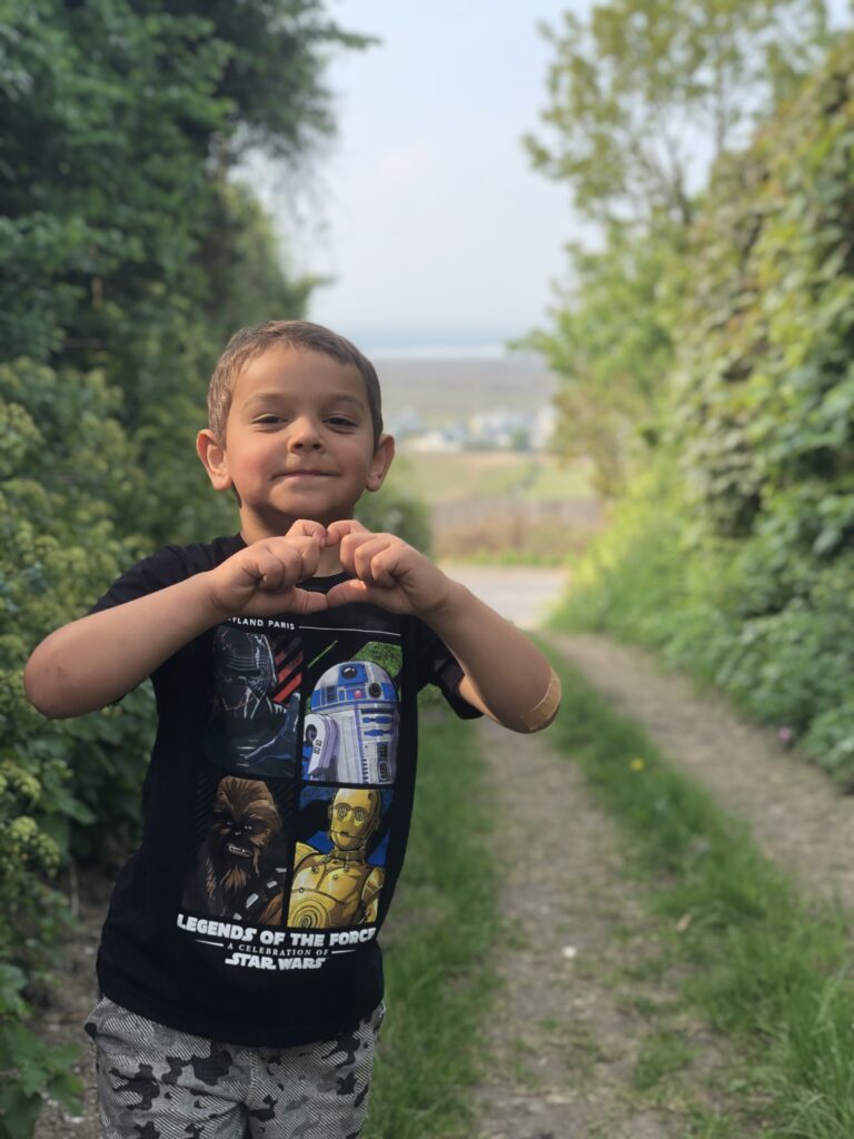 Young child poses his hand to form a heart whilst standing before trees outside