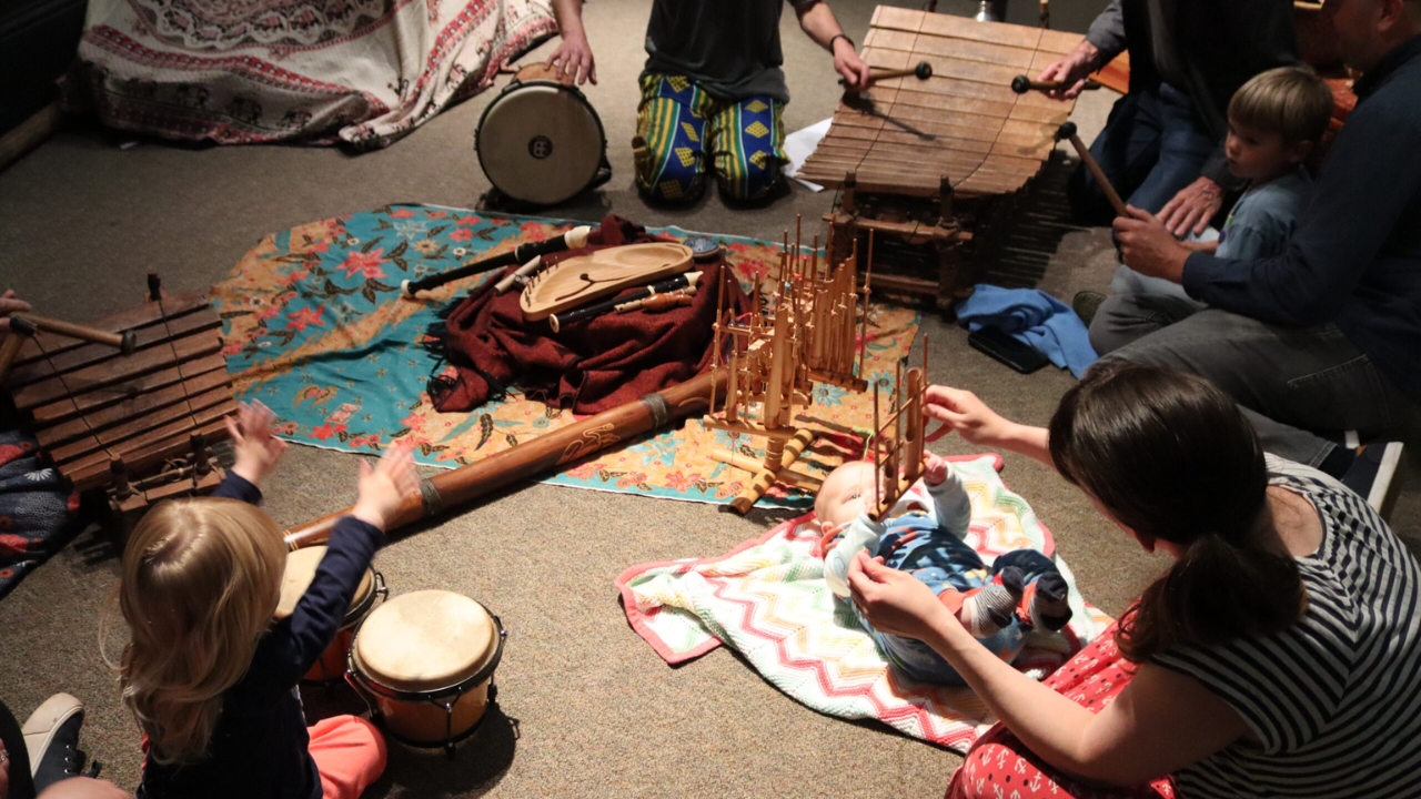 a mother and baby on the floor playing instruments, while a toddler to the left bangs on a drum
