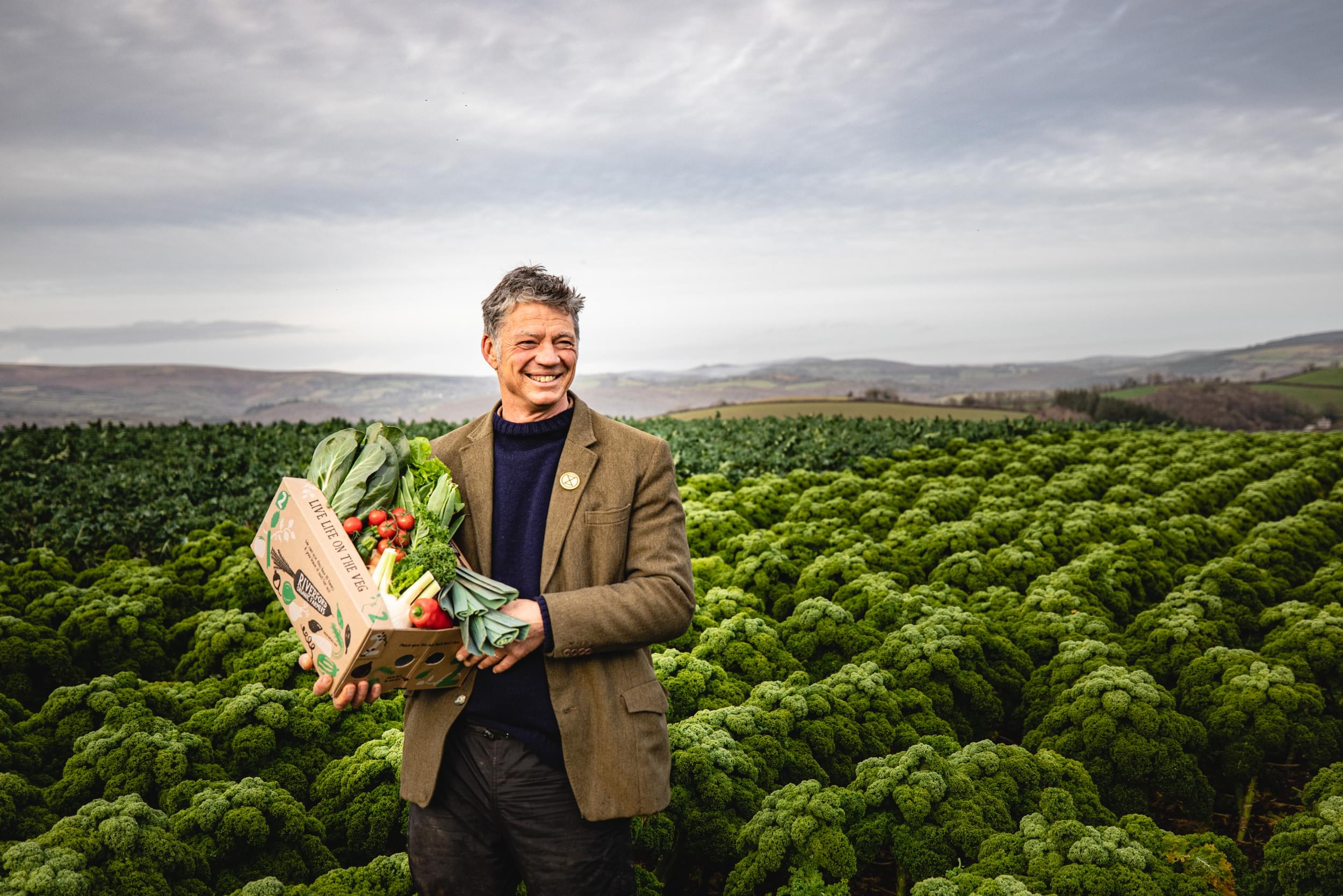 Guy, founder of Riverford, standing in a field holding a box of vegetables.