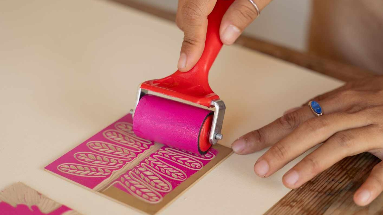 A pair of hands running a print roller covered with pink ink along a leaf shaped stencil