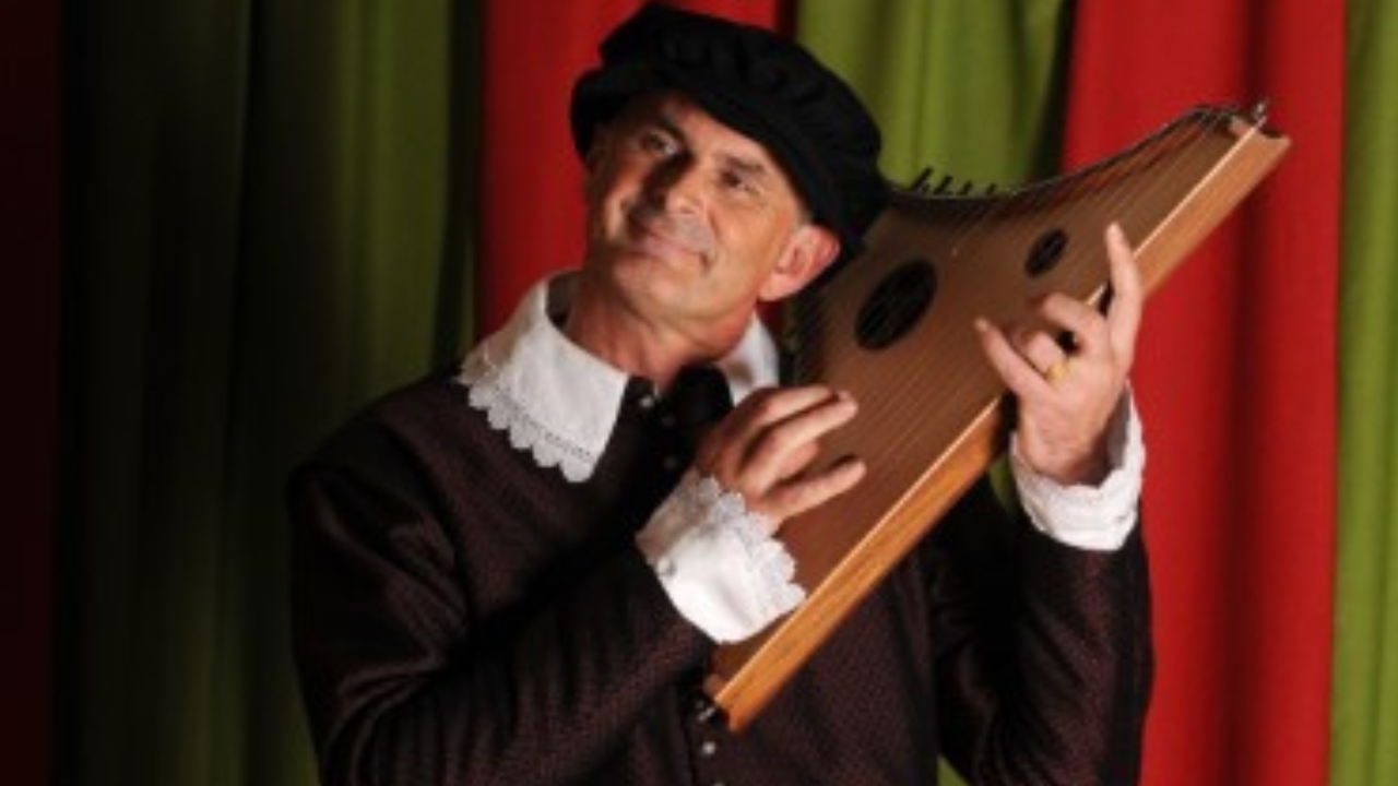Photo of the upper body of a middle aged man wearing a black Tudor tunic with a frilly white collar and a black cap, playing a Tudor string instrument.